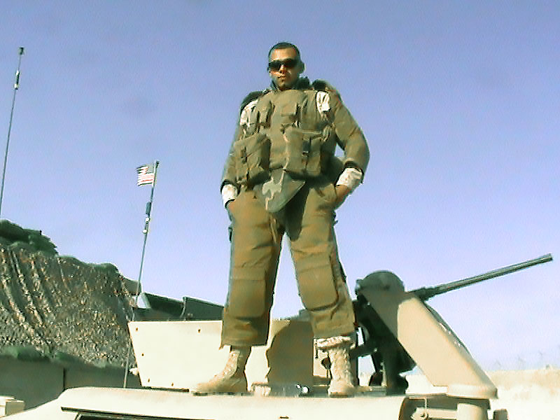Roberto in full tactical gear and sunglasses stands confidently atop a military vehicle, with an American flag in the background under a clear sky.
