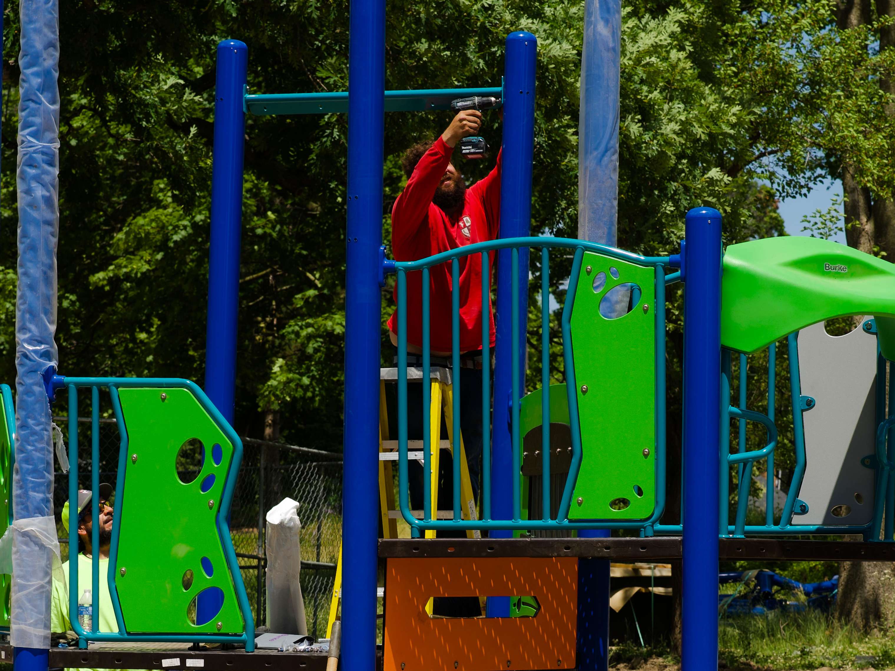 Tim stands on a ladder using a power drill to assemble a colorful playground structure surrounded by trees.