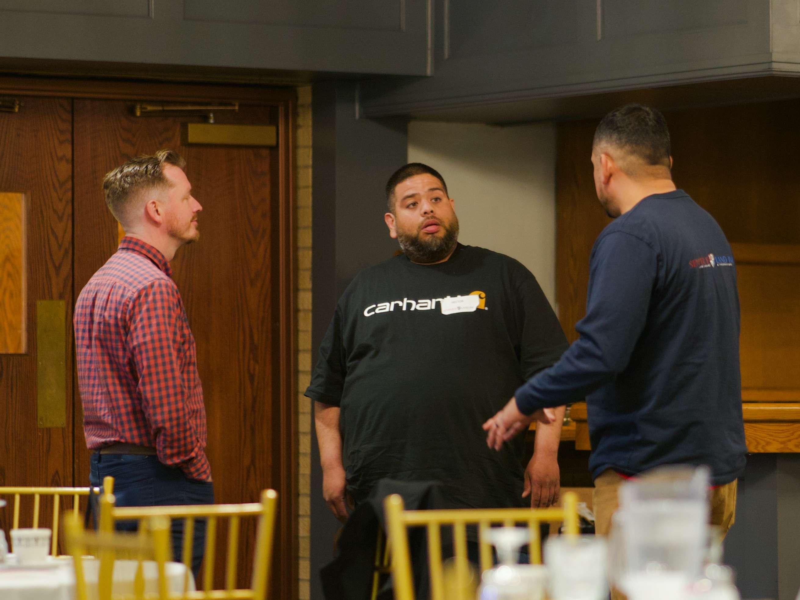 Three men have a conversation inside a banquet hall, standing near gold chairs and round tables, with one wearing a Semper Fi Land shirt.
