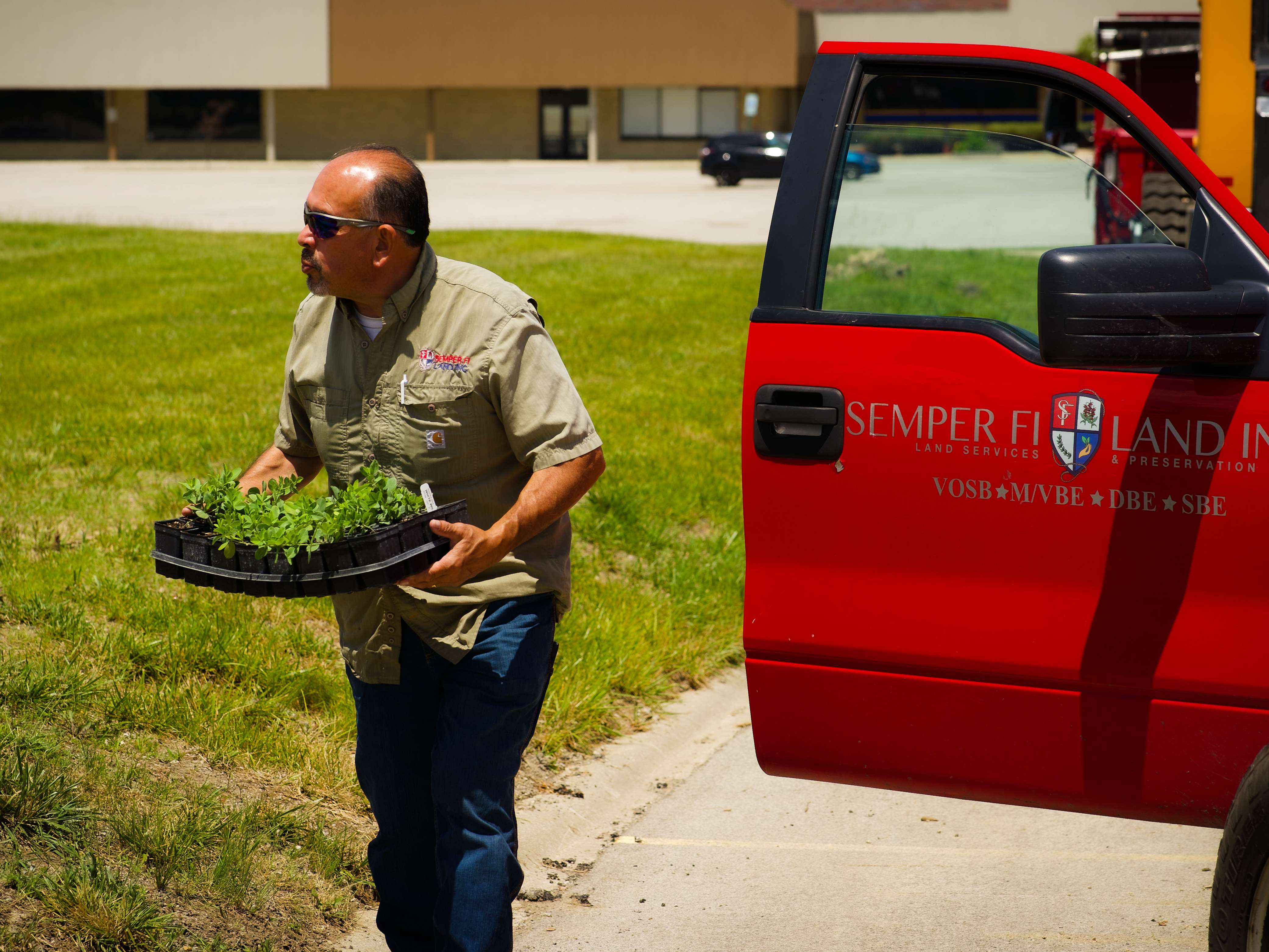 Rudy in a Semper Fi Land Services shirt carries a tray of young plants across a grassy area next to a red company truck.